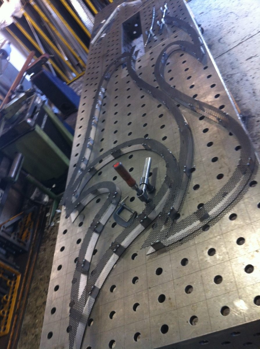 Curved perforated steel strips clamped to a fabrication table in a workshop, with machine tools alongside