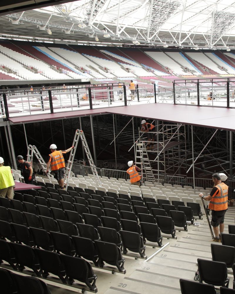 Crews in hard hats and hi-vis on ladders and scaffold, building a raised deck over the lower seating at the London Stadium