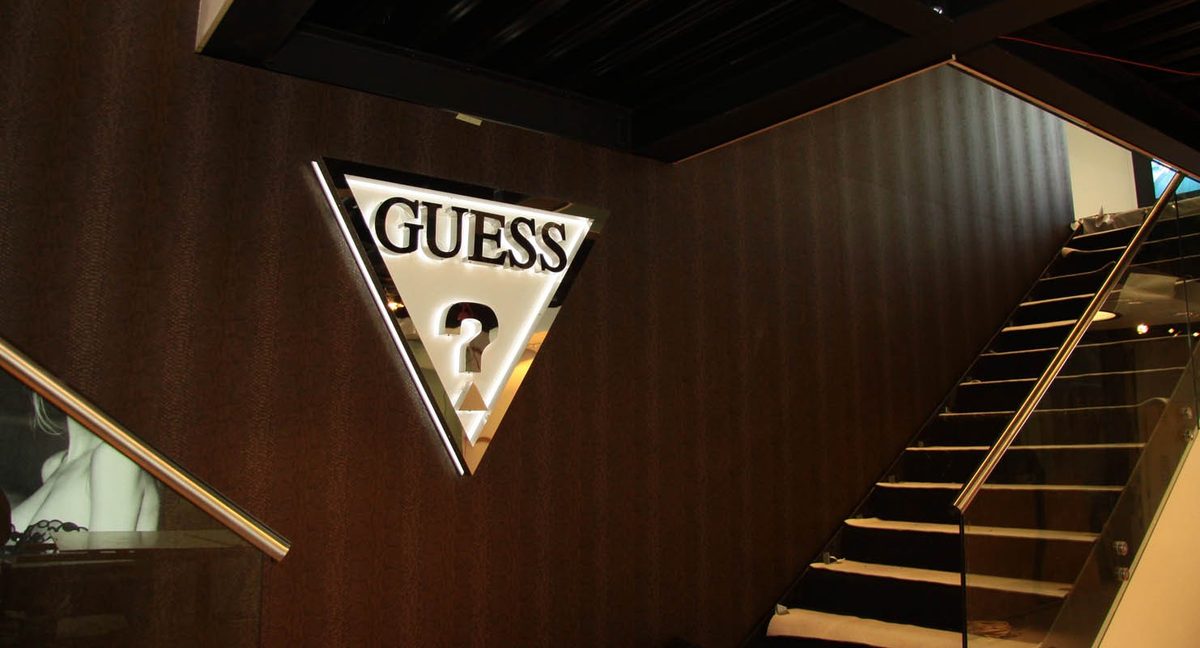 The finished Guess Jewellery showroom: an illuminated triangular Guess logo on a dark timber wall beside a glass-balustraded staircase